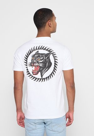 Homme portant un T-shirt blanc avec un motif tête de panthère noire rugissante dans un cadre circulaire ressemblant à des épines dans le dos, associé à un jean bleu clair.