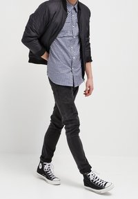 Svart bomberjacka över en blå och vit rutig skjorta, tillsammans med svarta skinny jeans och svarta höga sneakers.