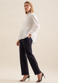 Weißes Hemd mit entspannter Passform, langen Ärmeln und seitlichen Schlitzen, kombiniert mit geraden, navyfarbenen Hosen und schwarzen High-Heels.