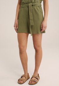 Olivově zelené strukturované šortky s ladícím opaskem, se volným střihem. Nošeny s hnědými sandály, které mají dvojité přezky a otevřenou špičku.