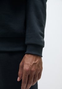 Sudadera gris oscuro con puños y dobladillo acanalados, que presenta un primer plano de una mano reposando sobre la tela, destacando la textura y los detalles de costura.