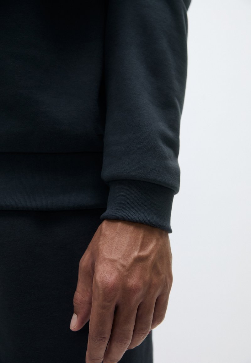 Sweatshirt gris foncé avec des poignets et un ourlet côtelés, mettant en avant un gros plan d'une main reposant sur le tissu, soulignant la texture et les détails de couture.