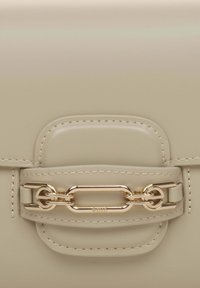 Beżowa skórzana torba o eleganckim designie, z złotym łańcuchem ozdobnym i detalami stębnowania na zapięciu klapy.