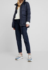 Marinblå dunjacka med dragkedja, hög krage och framfickor, i kombination med en krämfärgad tröja, mörka jeans och flerfärgade sneakers.