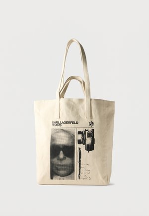Bež platnena torba z črnim grafičnim potiskom, ki prikazuje obraz z očali za sonce, fotoaparat, pero in besedilo "KARL LAGERFELD JEANS".