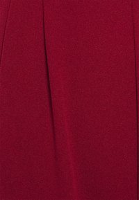 Tumši sarkans audums ar mīkstu, teksturētu virsmu, kurai ir smalka spīduma un krokotu dizaina detaļas, radot elegantu un izsmalcinātu izskatu.