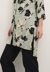 Tunique florale en vert clair avec des motifs de fleurs noirs et crème, texture côtelée et fentes sur les côtés. Associée à un jean noir.