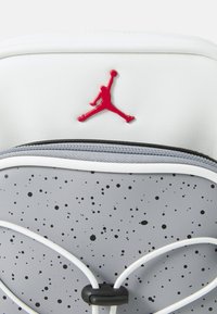 Zaino grigio e bianco con superficie testurizzata punteggiata di nero, logo Jumpman rosso e cinghie bianche a contrasto.