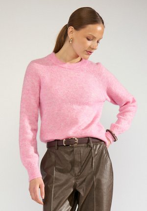 Femme regardant vers le bas, portant un pull rose duveteux, un pantalon en cuir marron avec une ceinture, des boucles d'oreilles créoles dorées et un bracelet à son poignet droit.