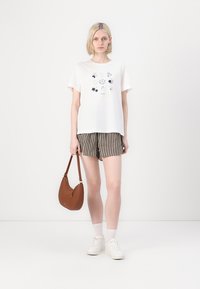 T-shirt blanc à manches courtes avec un imprimé graphique, accompagné d'un short rayé. Le modèle tient un sac à main marron et courbé. Il porte des baskets blanches et des chaussettes.