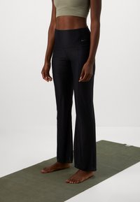 Cilvēks stāv basām kājām uz zaļas jogas paklāja, valkājot melnu platleņķa sporta bikses un olīvzaļu sporta krūšturi pret balta fona.