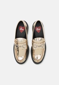 Mocasins de couro patente em ouro com uma sola preta, apresentando um detalhe em corrente em forma de coração prateado e um logótipo de coração vermelho na palmilha.