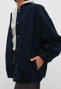 Osoba nosząca granatową kurtkę zapinaną na zatrzaski na jasnoszarym hoodie, z jedną ręką w kieszeni kurtki, na jednolitym tle.