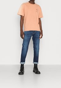 Broskvově oranžové tričko s krátkým rukávem a malým potiskem loga, kombinované s úzkými tmavě modrými džíny a černými šněrovacími botami s ohrnutými lemy.