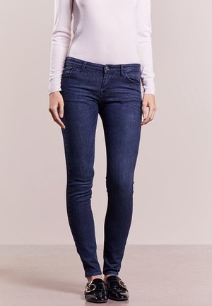 Femme portant un jean skinny bleu foncé, un haut blanc à manches longues et des chaussures slip-on noires avec un détail de boucle dorée, debout sur un sol neutre.