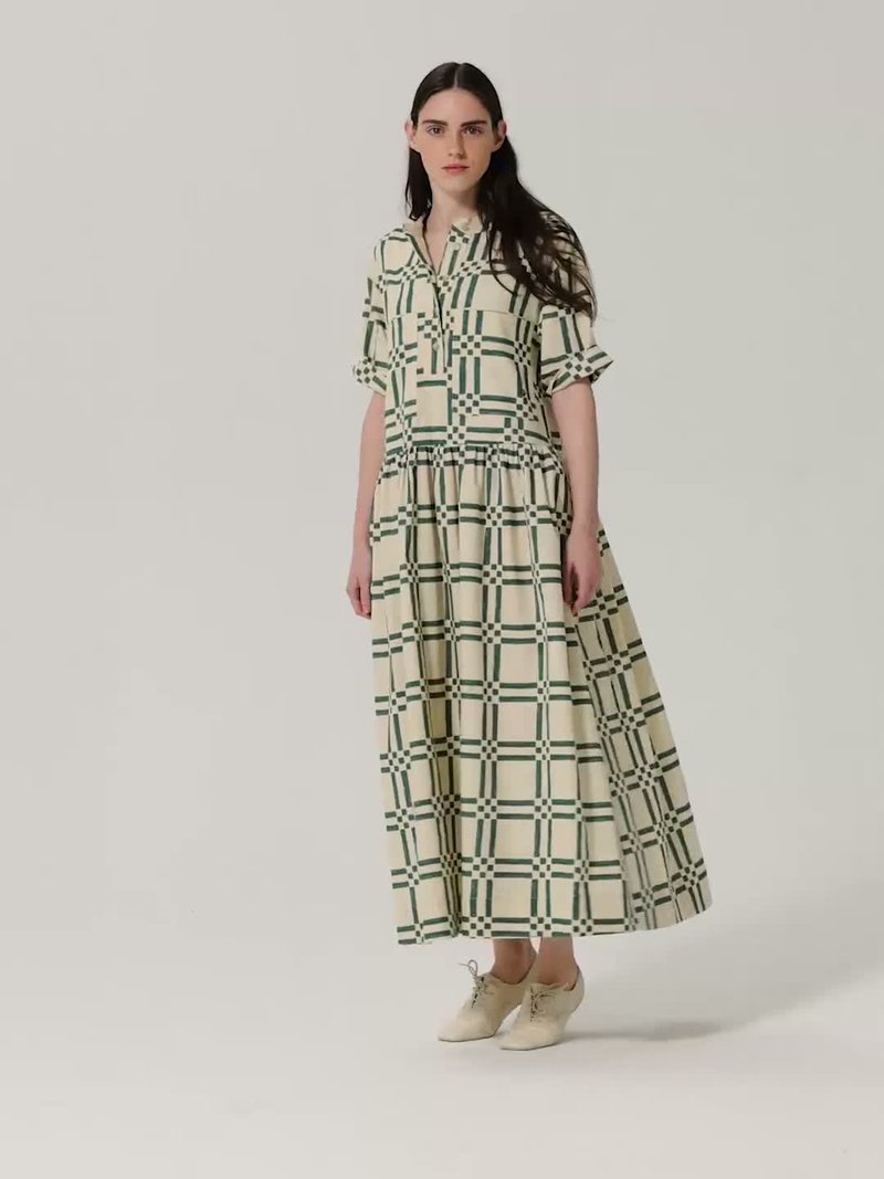 Femme aux longs cheveux foncés portant une longue robe à carreaux crème et verte avec manches retroussées et des chaussures beiges, se tenant devant un fond uni.
