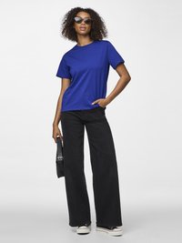 Žena s kovrčavom kosom nosi sunčane naočale mačjeg oblika, plavu majicu, crne široke hlače, tenisice i drži crnu torbu.