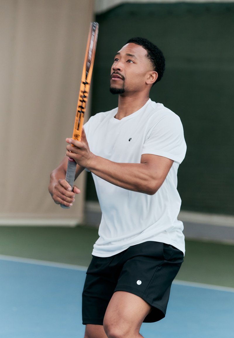 Tennis-spiller i en hvit kortermet skjorte og svarte shorts, som holder et oransje tennisracket med teksturert grep, innendørs på en bane.