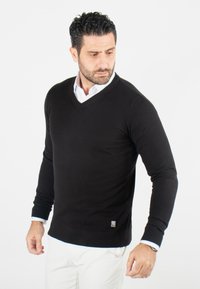 Pull noir en V fabriqué en tissu doux, avec des manches longues et des poignets côtelés. Présente un petit logo en accent sur l'ourlet.