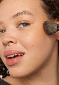 Aplikacja makijażu w toku, z użyciem miękkiego, płaskiego pędzla na jasnej karnacji. Cechy obejmują piercing w septum i subtelne piegi.