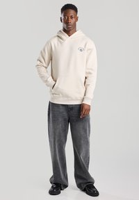 Sudadera beige con capucha y bolsillo frontal, que presenta un logo bordado en el pecho, combinada con pantalones de mezclilla gris de corte holgado y zapatos negros.