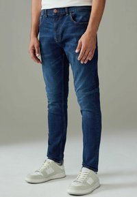 Jean bleu foncé en coupe slim avec un léger effet de décoloration, présentant un style cinq poches et une fermeture à bouton marron. Associé à des baskets blanches.