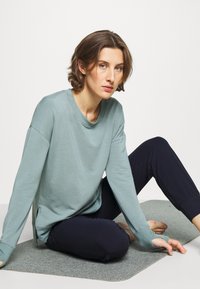 Helles türkisfarbenes Langarmshirt mit rundem Halsausschnitt und seitlichen Schlitzen, kombiniert mit enganliegenden dunkelblauen Hosen. Matte Textur, lässiges Design.