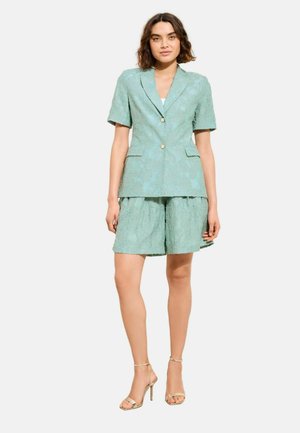 Nainen seisoo, pukeutuneena vaaleanvihreään, teksturoituun lyhythihaiseen bleiseriin ja siihen sopiviin leveälahkeisiin shortseihin sekä kultaisiin korkokenkäsandaaleihin.