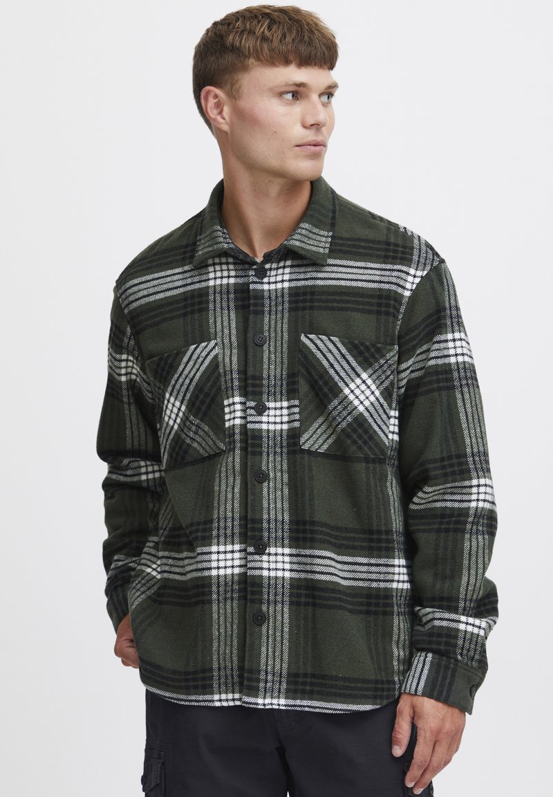 Cămașă din flanel verde cu carouri și dungi albe, cu două buzunare la piept, design cu nasturi și nasturi închiși la culoare pe o textură netedă.