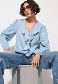 Blusa azul clara con frente con volantes, escote en V profundo y mangas abullonadas. Combinada con jeans de pierna ancha, sentada en una silla verde claro.