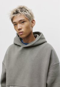 Light gray oversized hoodie with a front pocket and drawstring hood. Soft texture and relaxed fit. Model has short, spiky blonde hair.