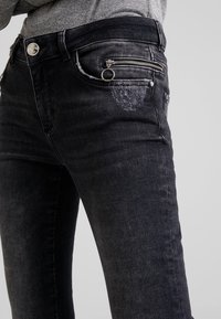 Personne portant un jean en denim noir délavé avec une poche à monnaie zippée et un anneau attaché à la tirette du zip sur le côté avant droit.