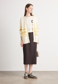 Cardigan crème avec des rayures jaunes, t-shirt blanc, jupe midi marron et baskets grises. Détail notable : logo brodé sur le t-shirt.