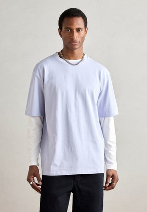 T-shirt court violet clair à manches courtes superposé sur une chemise blanche à manches longues, avec un col rond et une coupe décontractée. Un jean noir est porté.