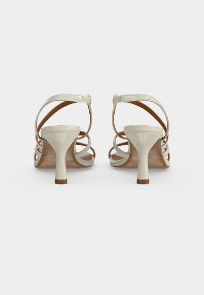 Paire de sandales blanches à talons avec fines brides et fermetures à la cheville, vues de dos sur un fond clair uni.