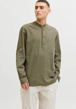 Uomo che indossa una camicia verde oliva a maniche lunghe con collo alla coreana abbottonato e pantaloni bianchi larghi, in posa con una mano in tasca.