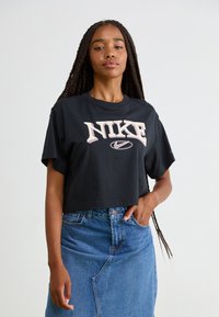 Vrouw met lange vlechten, gekleed in een zwart Nike T-shirt met korte mouwen en een blauwe spijkerrok, met één hand in de zak, tegen een effen lichte achtergrond.