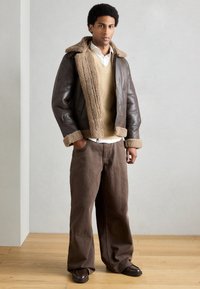 Barna bőrdzseki világosbarna műszőrme béléssel és mandzsettával. Bézs kötött mellény fehér ing felett. Barna, széles szárú nadrág és sötét cipő.