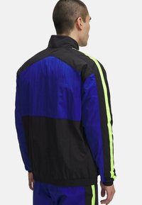 Herenjas in zwart, blauw en neongeel. Heeft een hoge kraag, een kleurblokontwerp en elastische manchetten, gemaakt van lichtgewicht synthetisch materiaal.