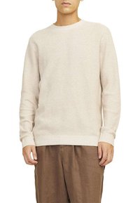 Pull en tricot beige clair avec un col rond, des poignets et un ourlet à côtes. Porté avec un pantalon en lin marron, présentant un tissu texturé.