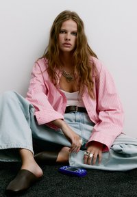 Camicia oversized a righe rosa, canotta bianca, jeans svasati azzurri chiari, mules marroni, anelli argentati e un dispositivo portatile blu su un tappeto scuro.