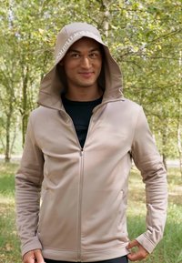 Šviesiai beige spalvos zip-up džemperis su dideliu gobtuvu ir reguliuojamomis raištelėmis, pasižymintis lygiu paviršiumi ir kontrastinga juoda vidine dalimi.