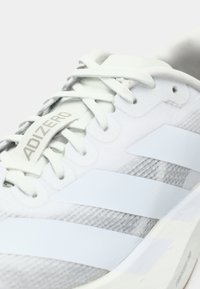 Viegls skriešanas apavs baltā krāsā ar gaiši pelēkiem akcentiem, kas izceļas ar trim spilgtām baltām svītrām, teksturētu augšpusi un "ADIZERO" logotipu.