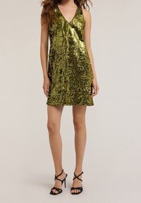 Zielona sukienka z cekinami, z dekoltem w kształcie litery V, bez rękawów i lekko rozkloszowanym dołem. Połączona z czarnymi sandałami na wysokim obcasie z rzemykami.