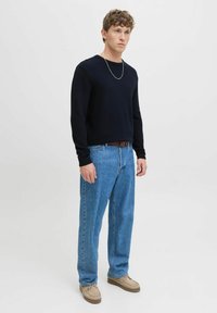 Pull en maille côtelée bleu marine, jean droit bleu clair, chaussures beiges. Un homme se tient contre un fond blanc uni avec une expression neutre.