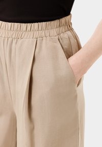 Beige kalhoty s elastickým pasem, které mají volné sklady a boční kapsy. Materiál má hladkou texturu a jemný lesk.