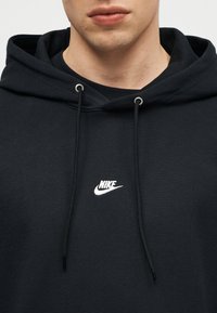 Κοντινό πλάνο ενός ατόμου που φοράει μαύρο φούτερ Nike με κορδόνια και μικρό λευκό λογότυπο Nike στο στήθος.