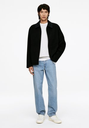 Schwarze Wollmischjacke mit kragenförmigem Ausschnitt, ausgestattet mit zwei Vordertaschen; kombiniert mit hellblauen geraden Jeans und weißen Sneakers.