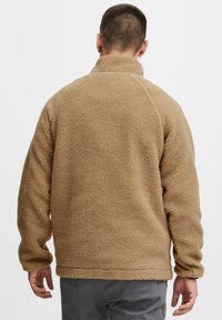 Chaqueta de forro polar color marrón claro con cuello alto, textura suave, mangas largas y hombros sin costuras, diseñada para proporcionar calor y comodidad.