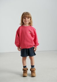 Różowa bluza z napisem "American Vintage", granatowe szorty oraz beżowe buty z pluszowymi wstawkami. Tło jest jasnoszare i minimalistyczne.